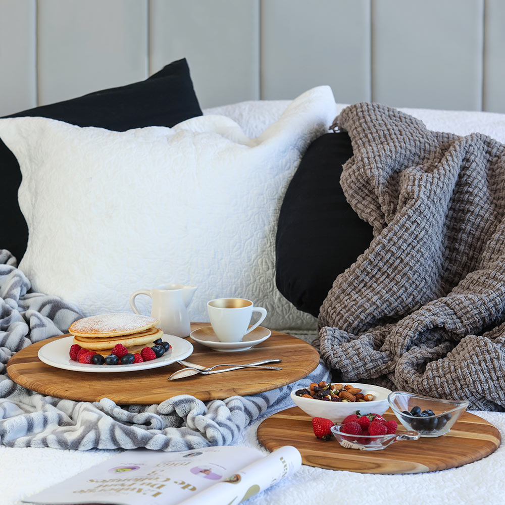 Edge grain round teak boards serving pancakes and fruit for a cozy breakfast in bed among pillows and blankets.