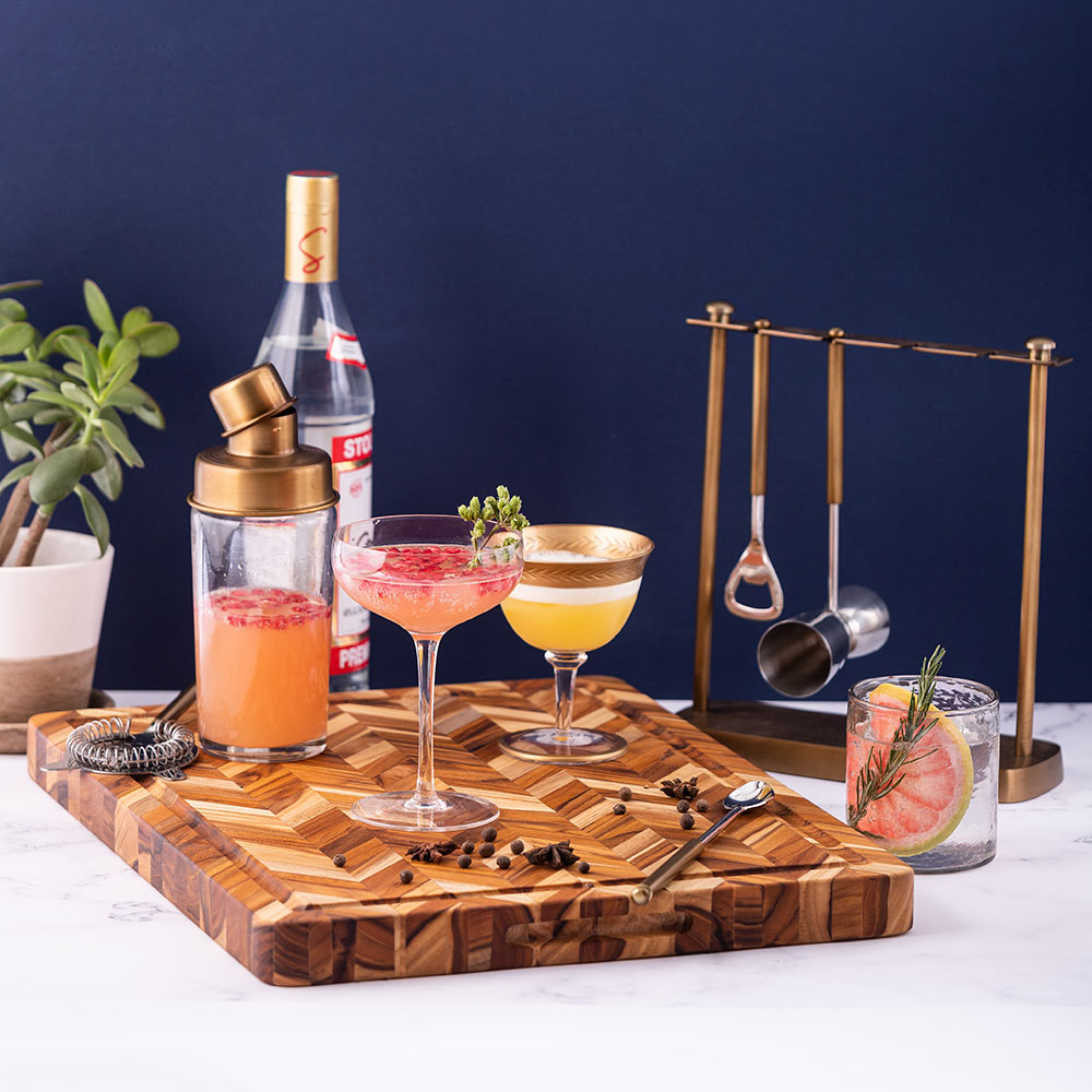 Teak herringbone cutting board with juice canal, styled as a bar station with cocktails and gold tools.