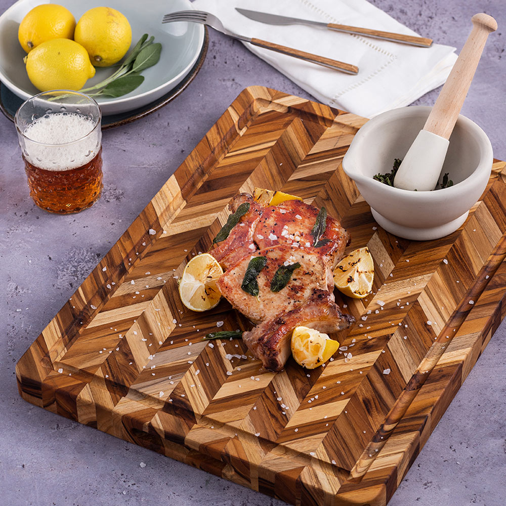 Large teak herringbone cutting board with a juice canal, featuring cooked pork chops and lemons.