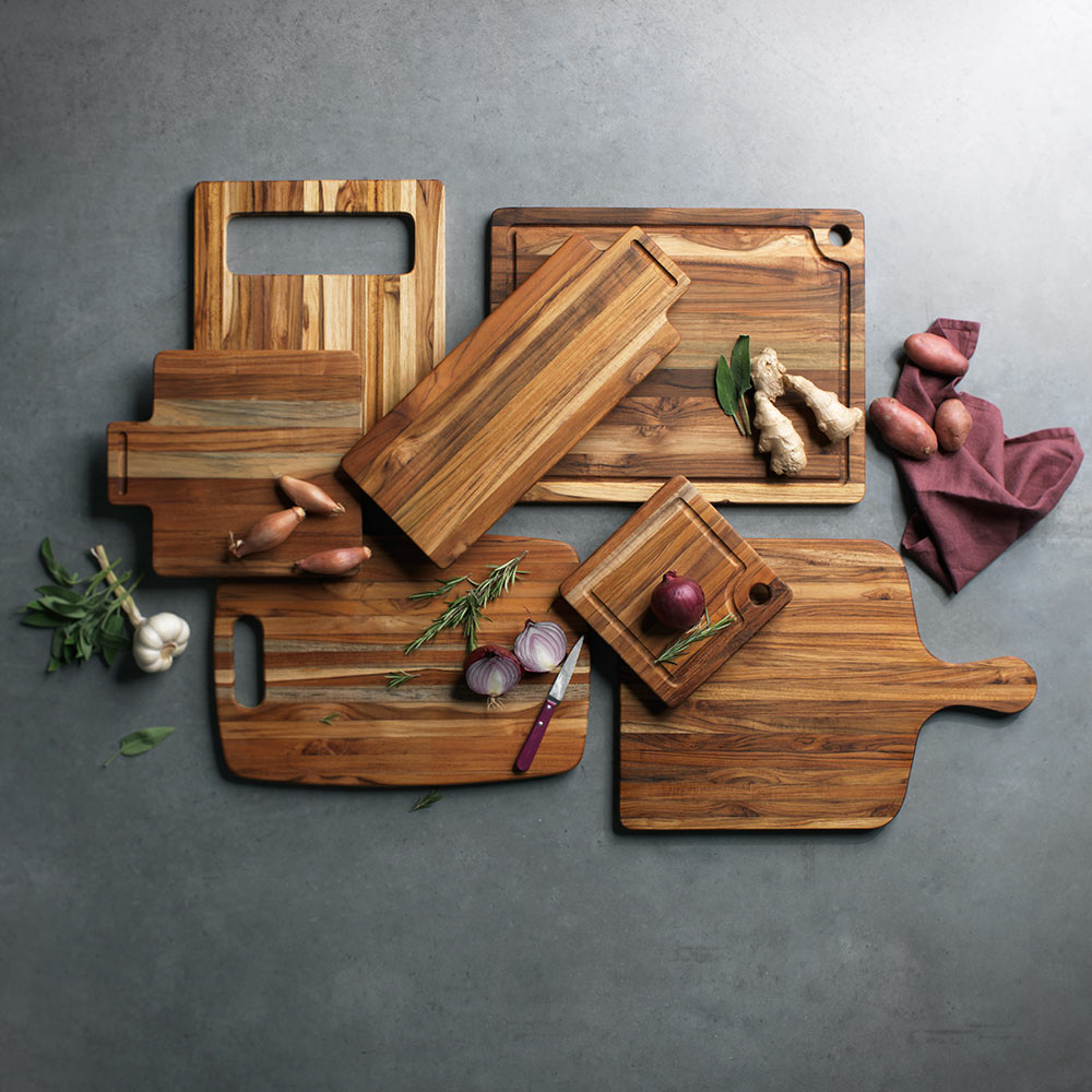 Various teak wood cutting and serving boards arranged on a grey surface with fresh vegetables and herbs.
