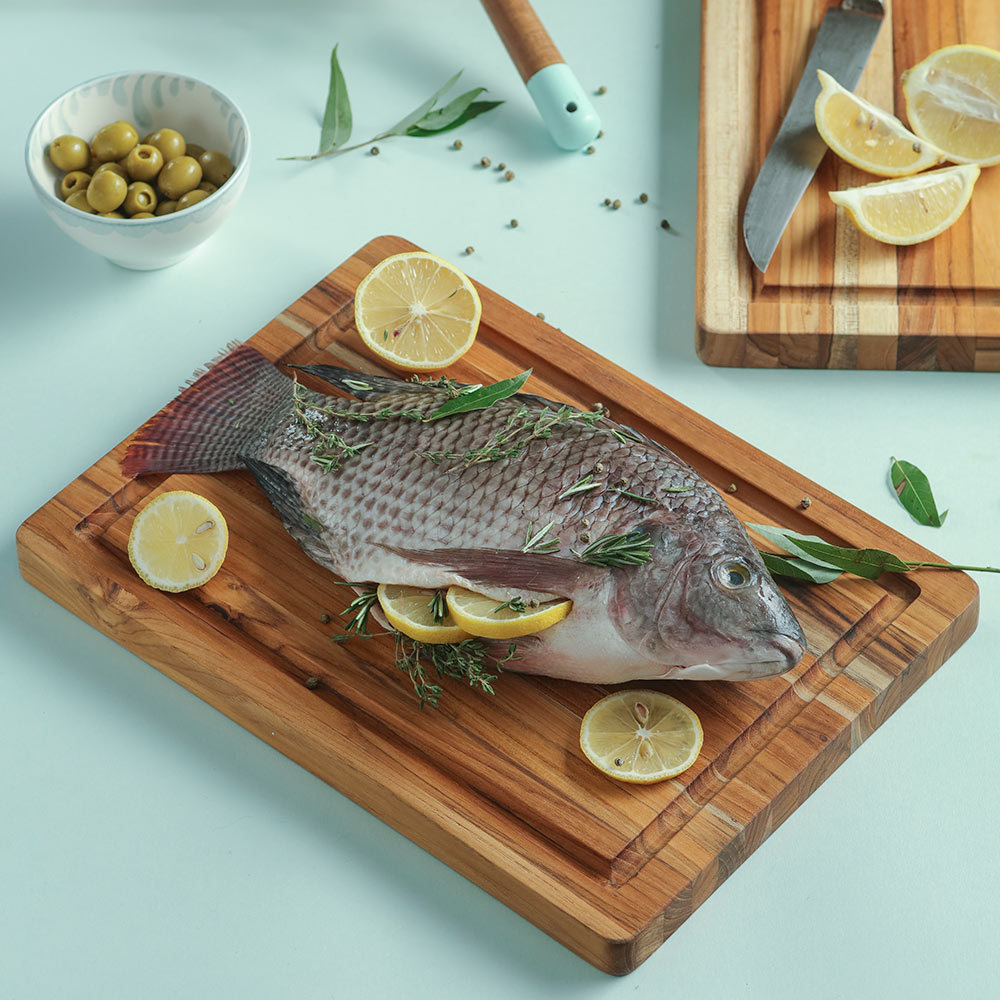 Thin teak edge grain cutting board with a juice groove, featuring a whole fish, herbs, and fresh lemon slices.