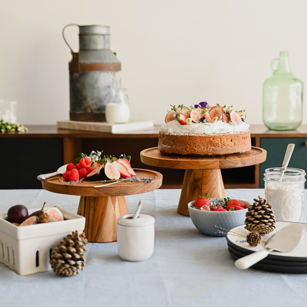 Traditional teakwood cake stand used as an elevated centerpiece for a fruit-topped cake on a rustic table.