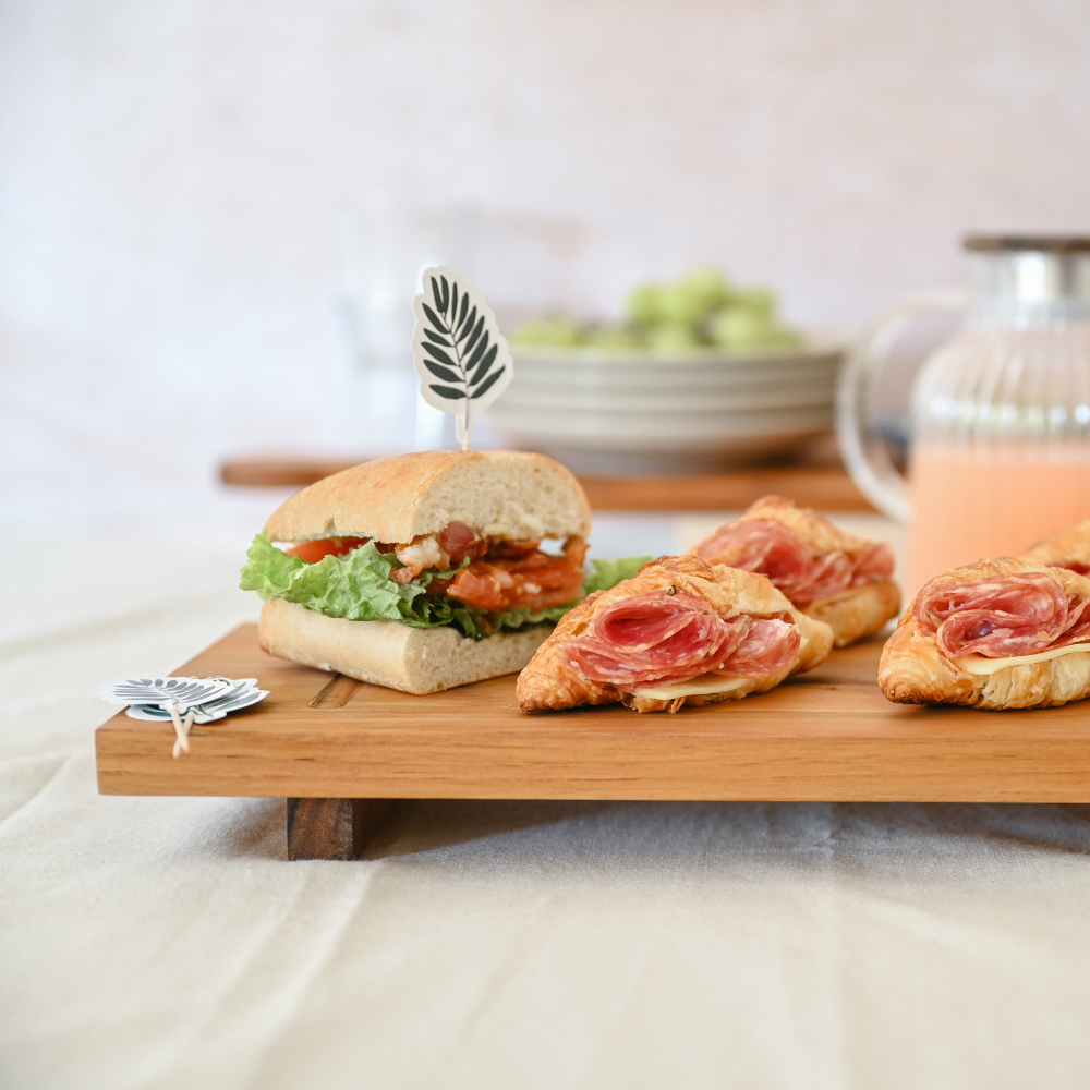 Small elevated teakwood food stand displaying savory sandwiches and croissants on a neutral tablecloth.