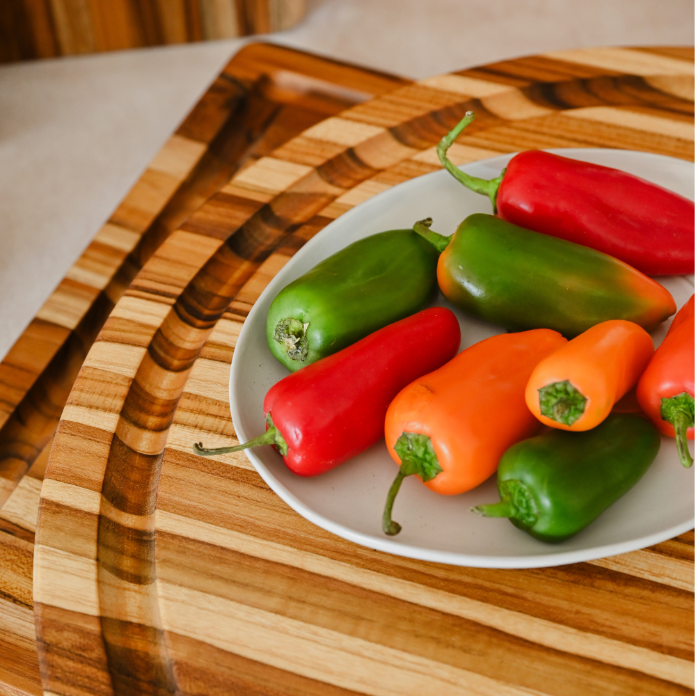 Round teakwood carving board with juice groove and a plate of colorful fresh peppers.