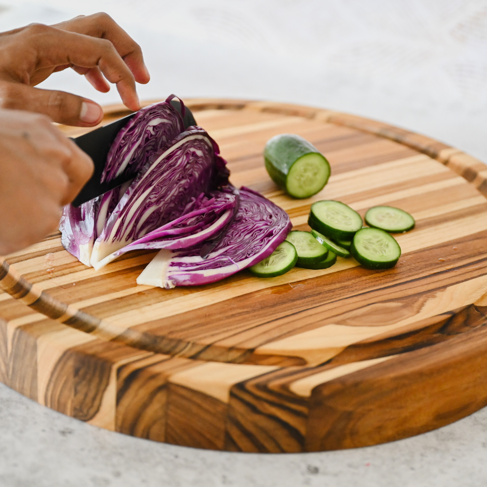Round edge grain teakwood carving board used to slice red cabbage and cucumbers with a black knife.