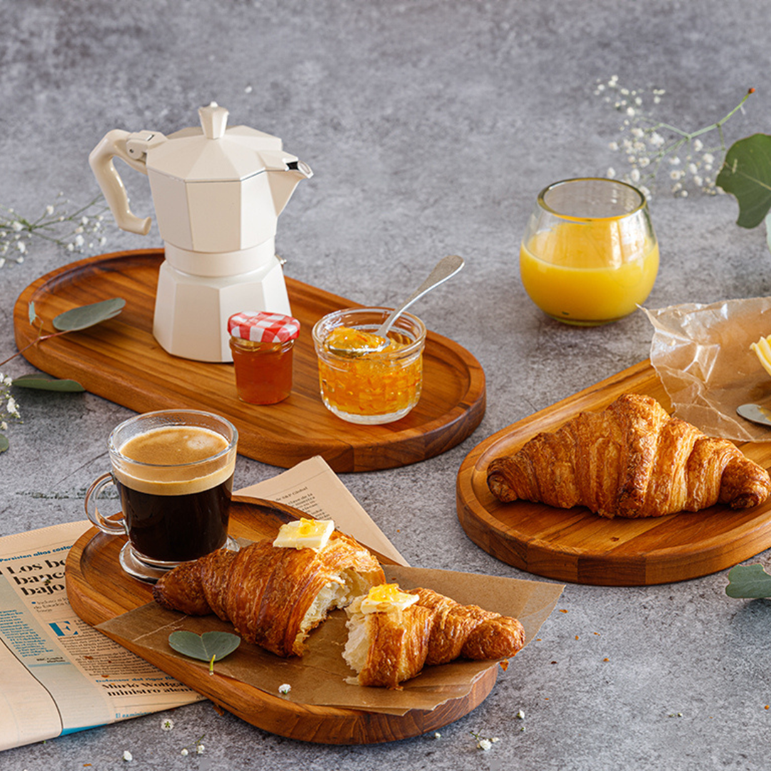 Essential oval teak wood serving trays styled with croissants, a white moka pot, coffee, jam, and orange juice on a textured tabletop.
