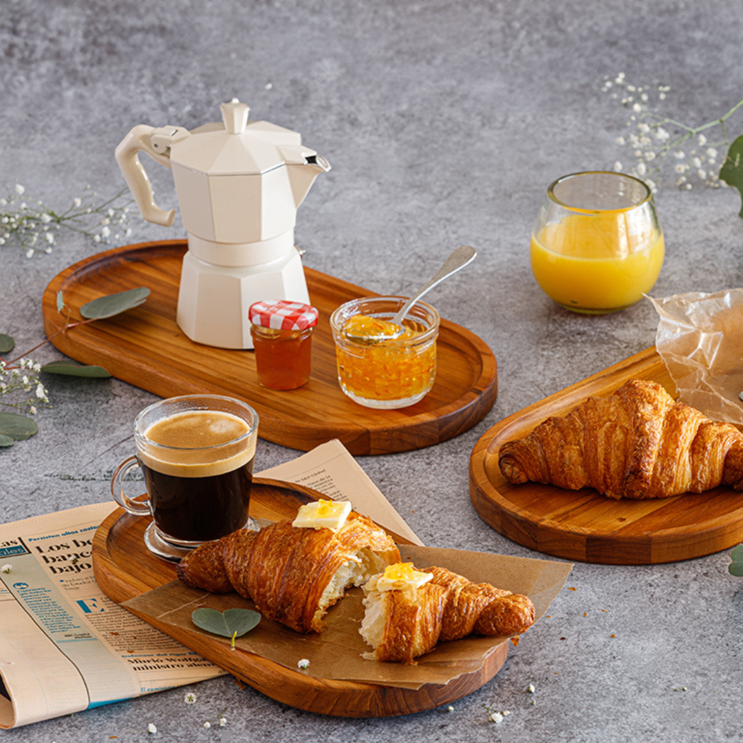 Medium oval teakwood serving trays styled with breakfast items including croissants, coffee, and jam on a grey surface.
