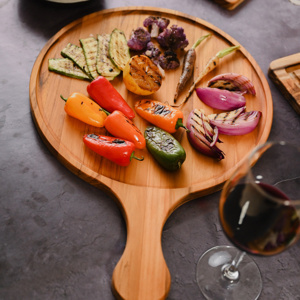 Round teakwood paddle board featuring grilled vegetables and colorful peppers on a dark, textured table.