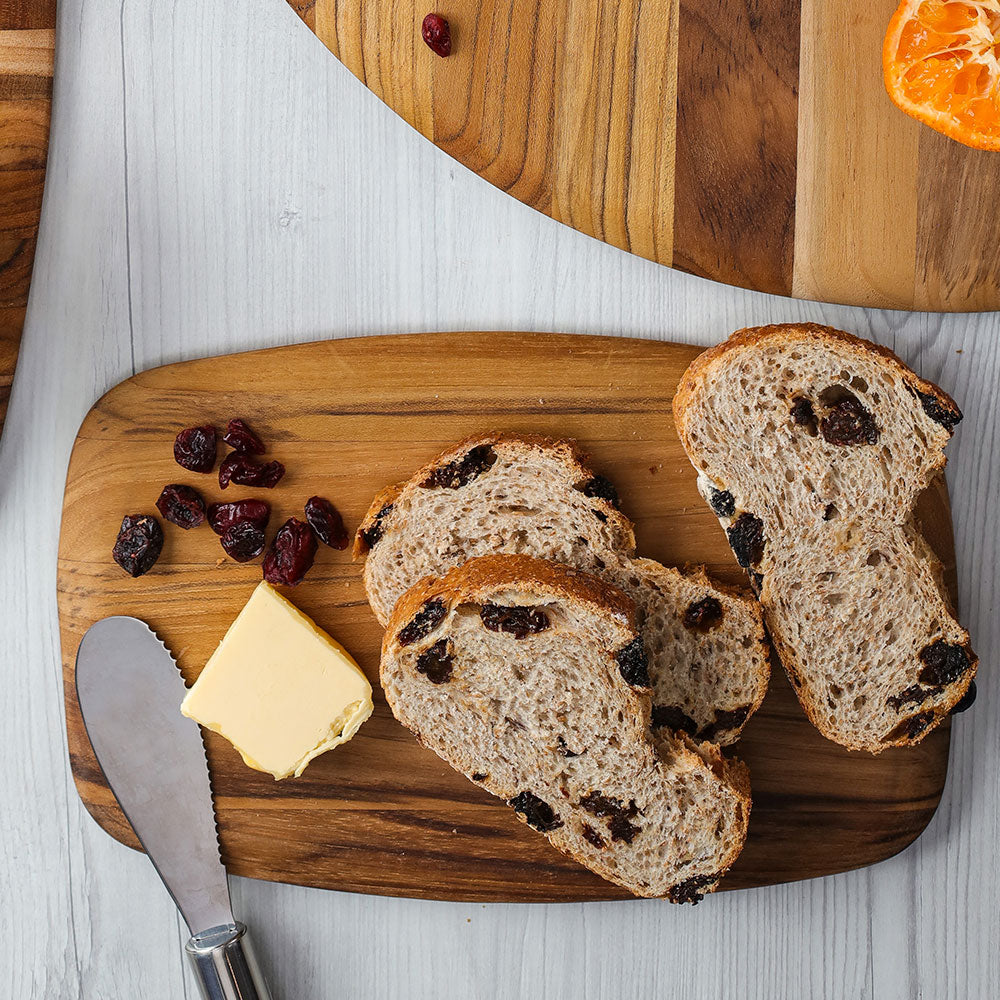 Sustainable teak serving board with rounded edges holding sliced raisin bread and butter for a healthy breakfast.