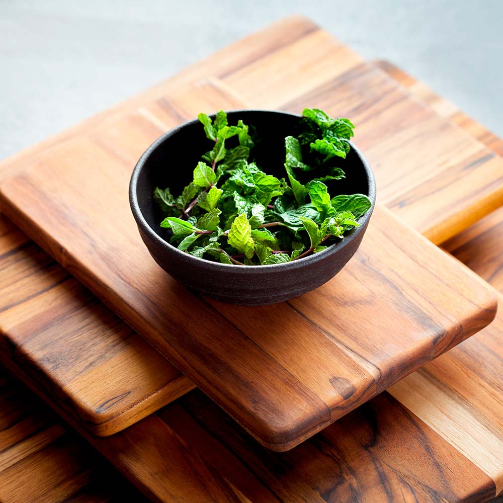 Rectangular edge grain teak cutting boards stacked with a black bowl of fresh mint.