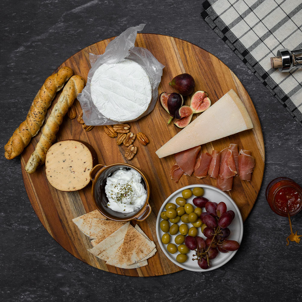 Round teakwood cutting board styled as a charcuterie platter with assorted cheeses, fruits, and olives.