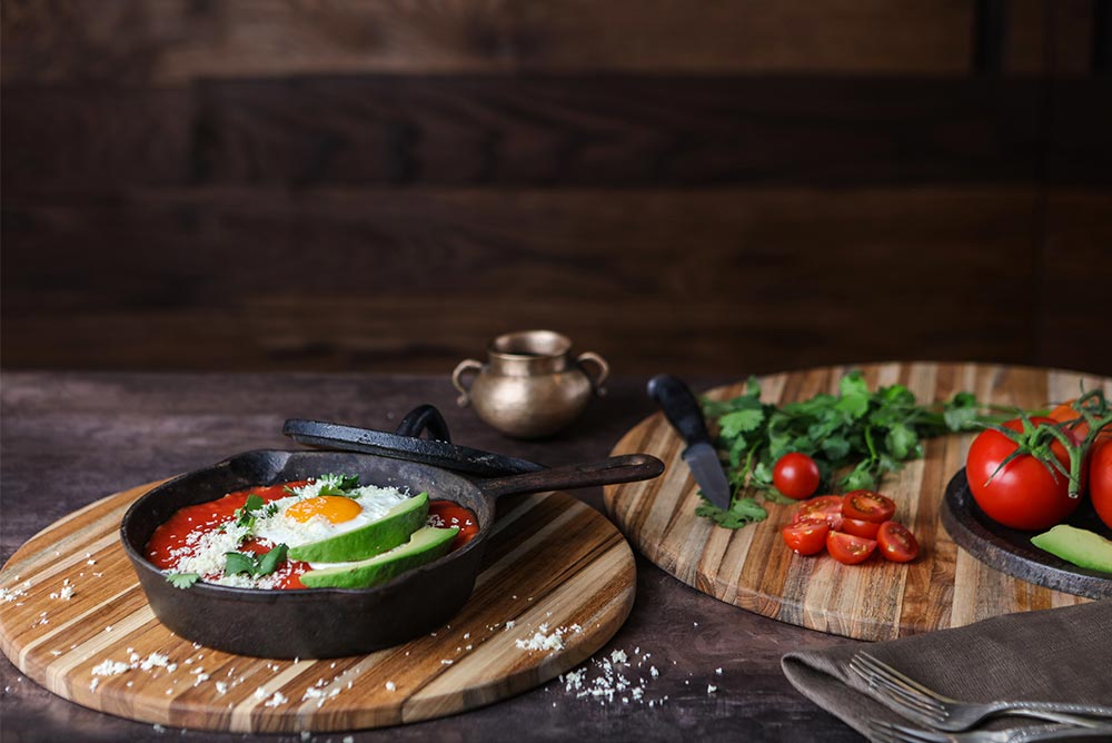 Round teakwood cutting boards with edge grain patterns serving a cast iron skillet meal and fresh vegetables.