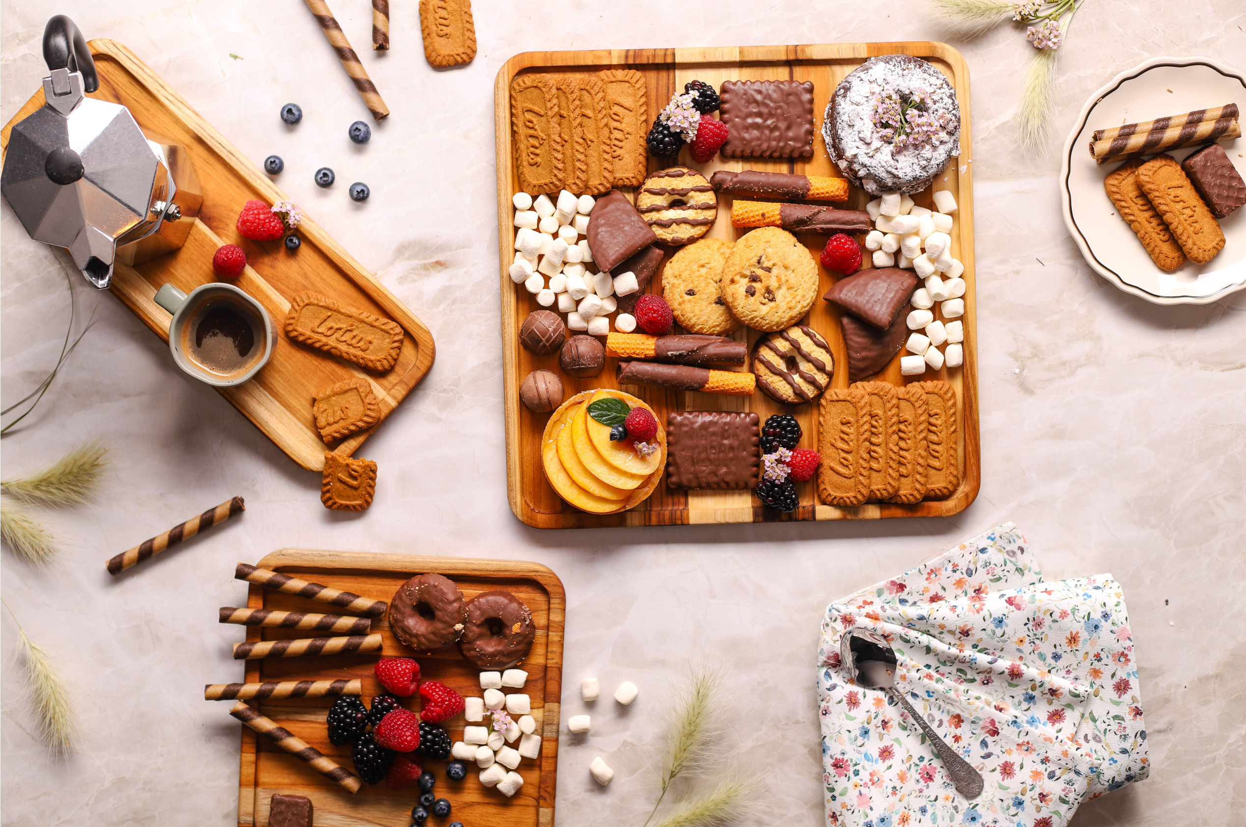 Square teakwood serving trays arranged with colorful desserts and fresh fruit on a light marble surface.